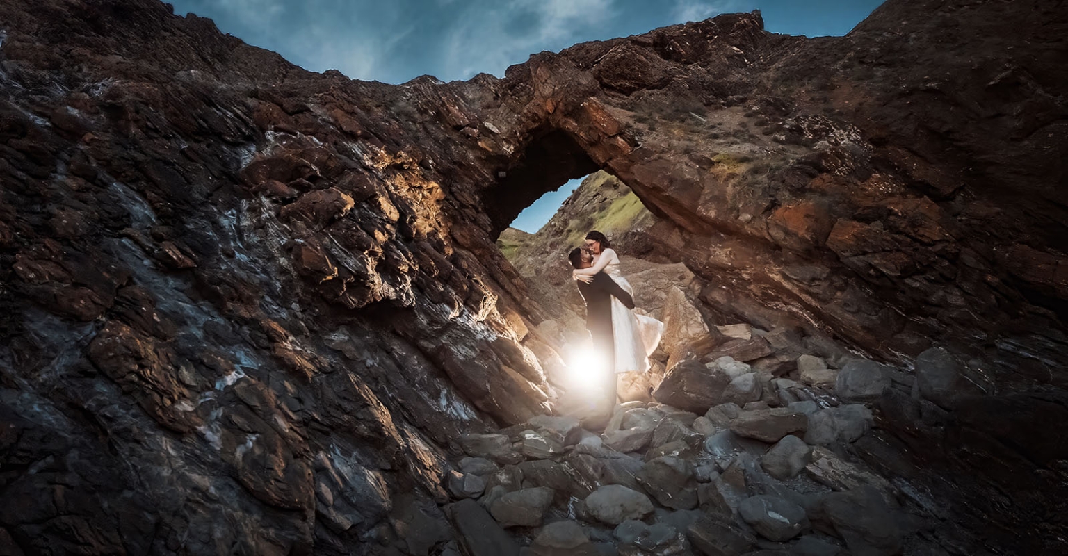 Ebony and Punit embracing beneath a natural rock arch at Second Valley, with sunlight glowing behind them