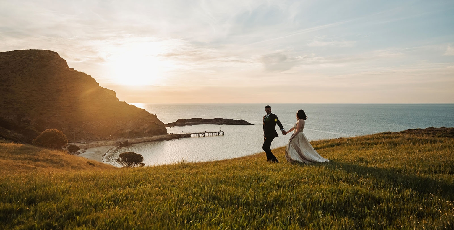 Ebony and Punit holding hands on a grassy coastal hill above Second Valley at sunset, with the bay and jetty below.