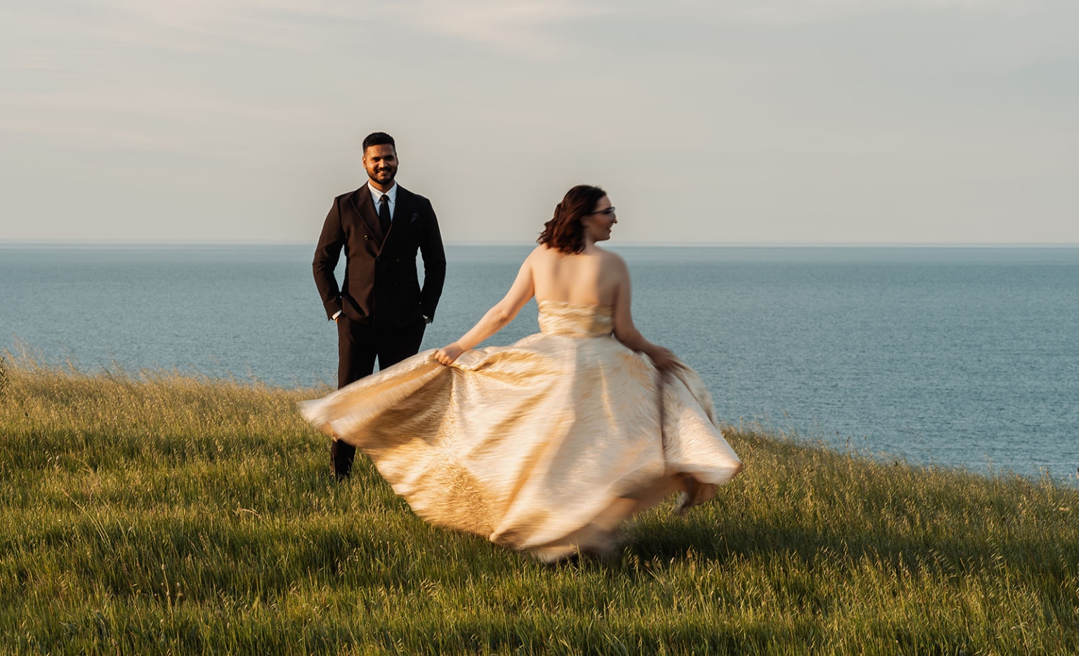 Ebony twirling her wedding dress on a grassy coastal hillside as Punit stands behind her overlooking the sea at Second Valley.