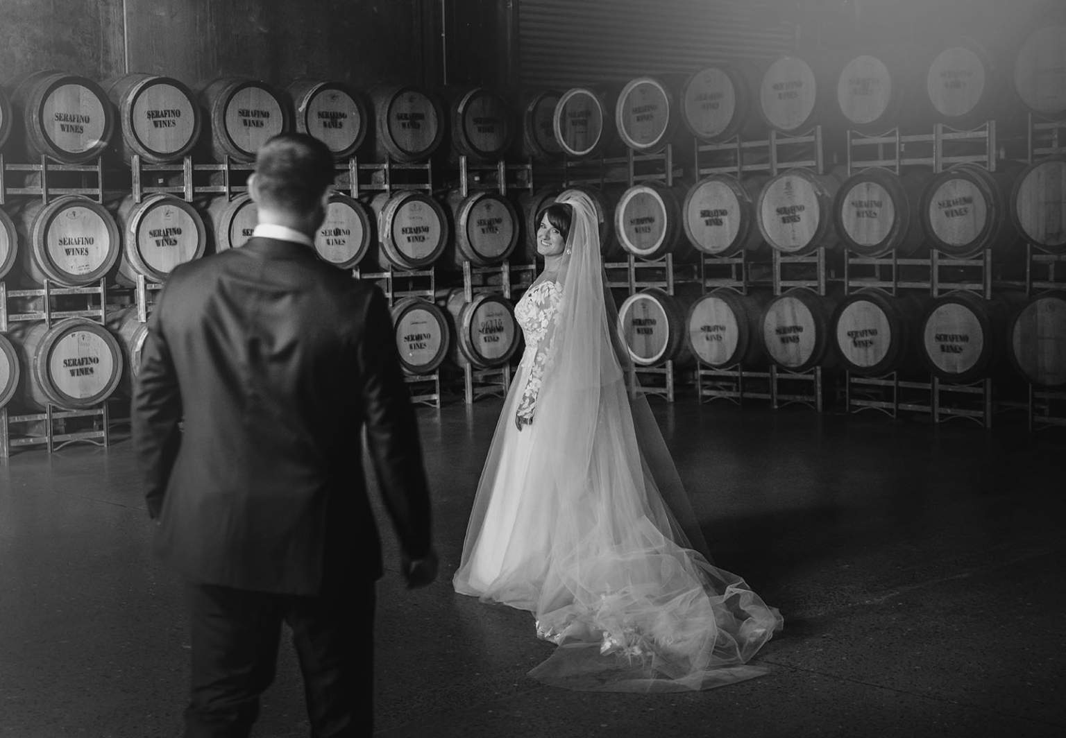 serafino winery wedding – maor & sasha 54 Bride in a lace wedding gown and long veil looks towards the groom among Serafino Wines barrels.