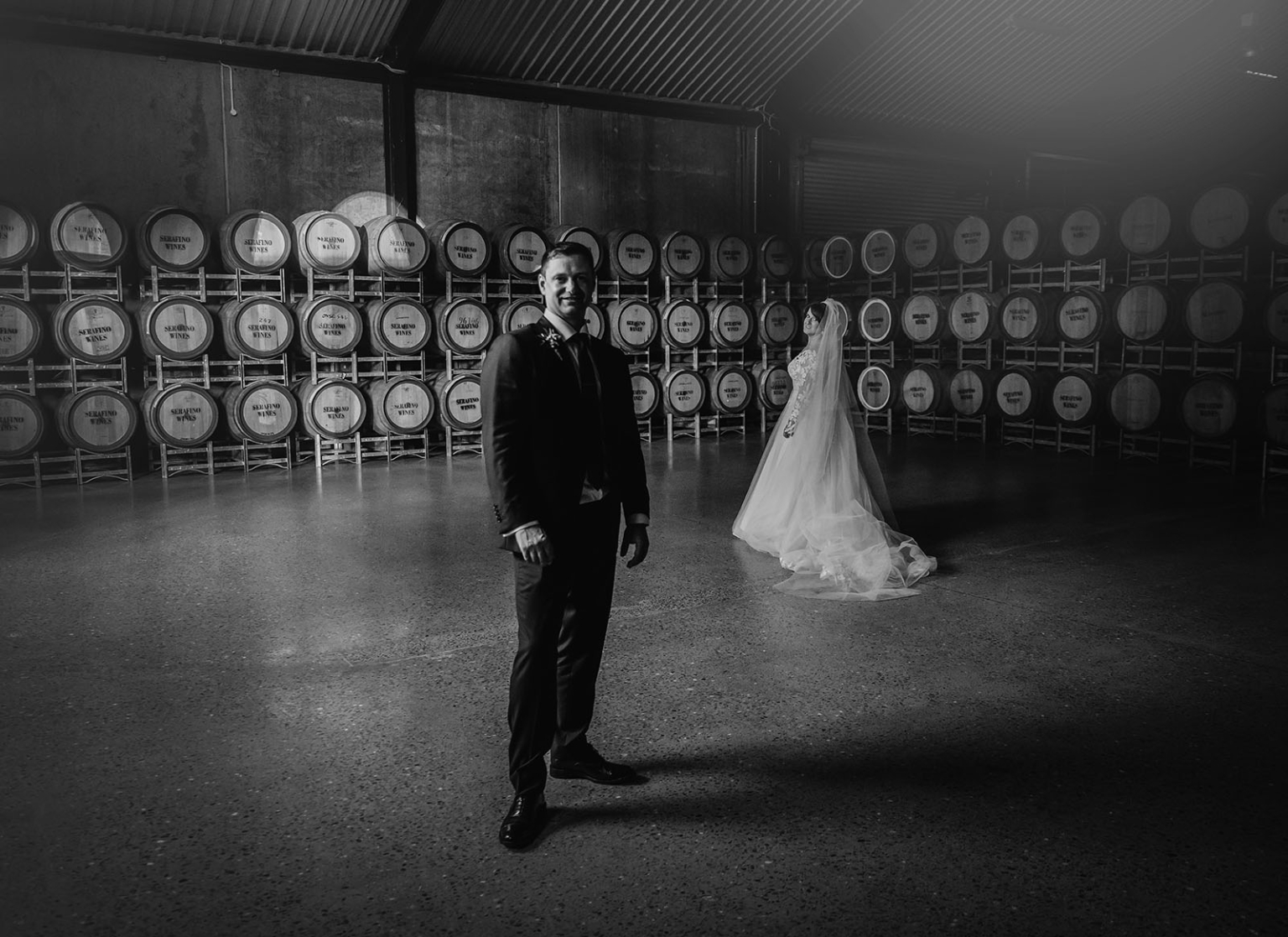 serafino winery wedding – maor & sasha 53 Groom in a dark suit and bride in a white gown standing among wine barrels in the Serafino Winery barrel room