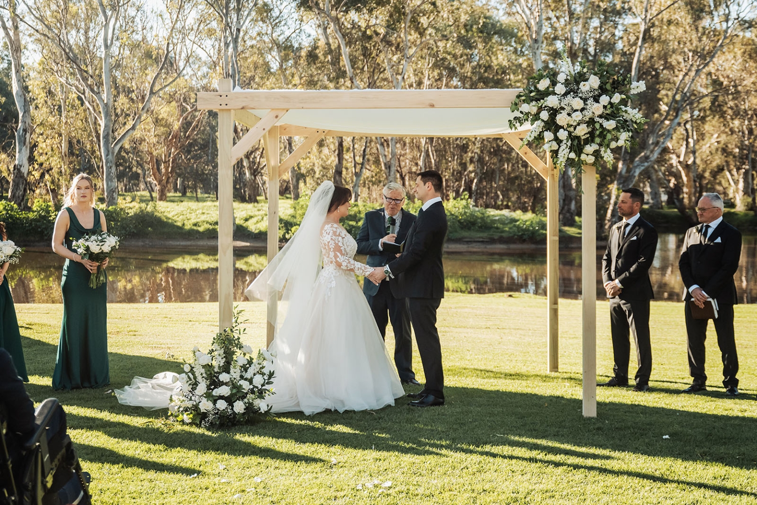 serafino winery wedding – maor & sasha 31 Couple holding hands beneath a timber floral arbour during a lakeside wedding ceremony at Serafino Winery