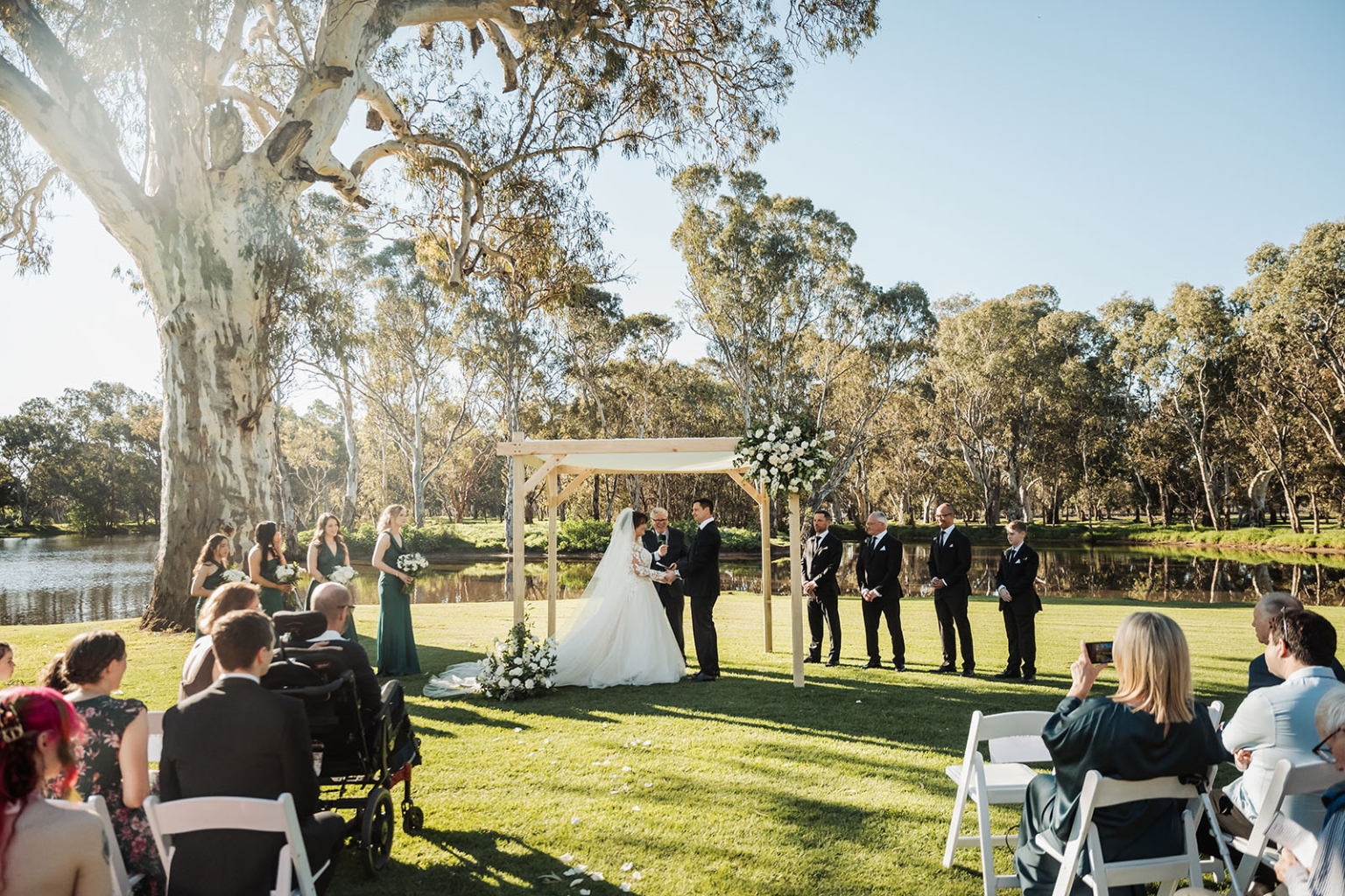 serafino winery wedding – maor & sasha 26 Outdoor wedding ceremony beneath a floral timber arbour beside a lake at Serafino Winery, with the wedding party and guests seated on the lawn.