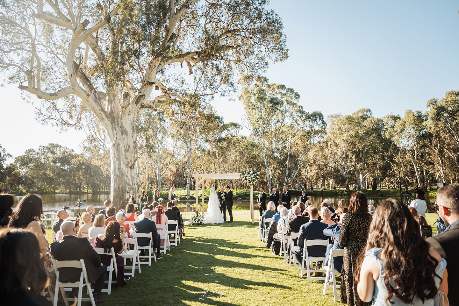 serafino winery wedding – maor & sasha 33 Wedding guests seated on a lawn beside a lake as the couple exchange vows beneath a floral arbour at Serafino Winery, McLaren Vale.