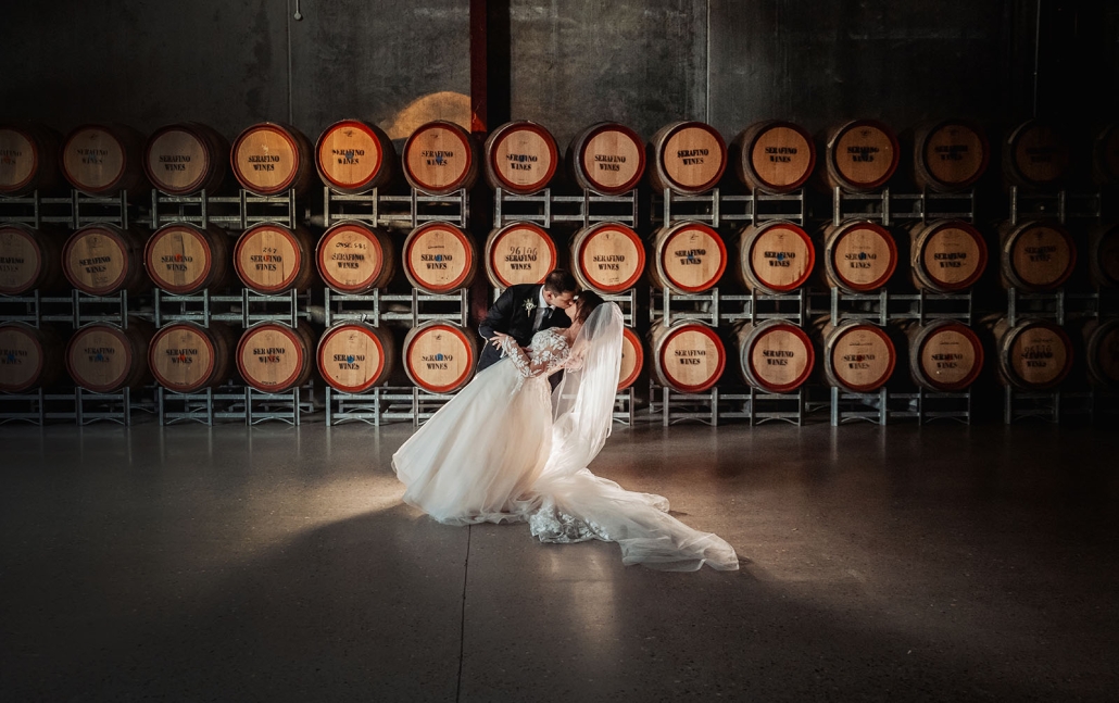 Bride and groom kissing in front of stacked Serafino Wines barrels at Serafino Winery