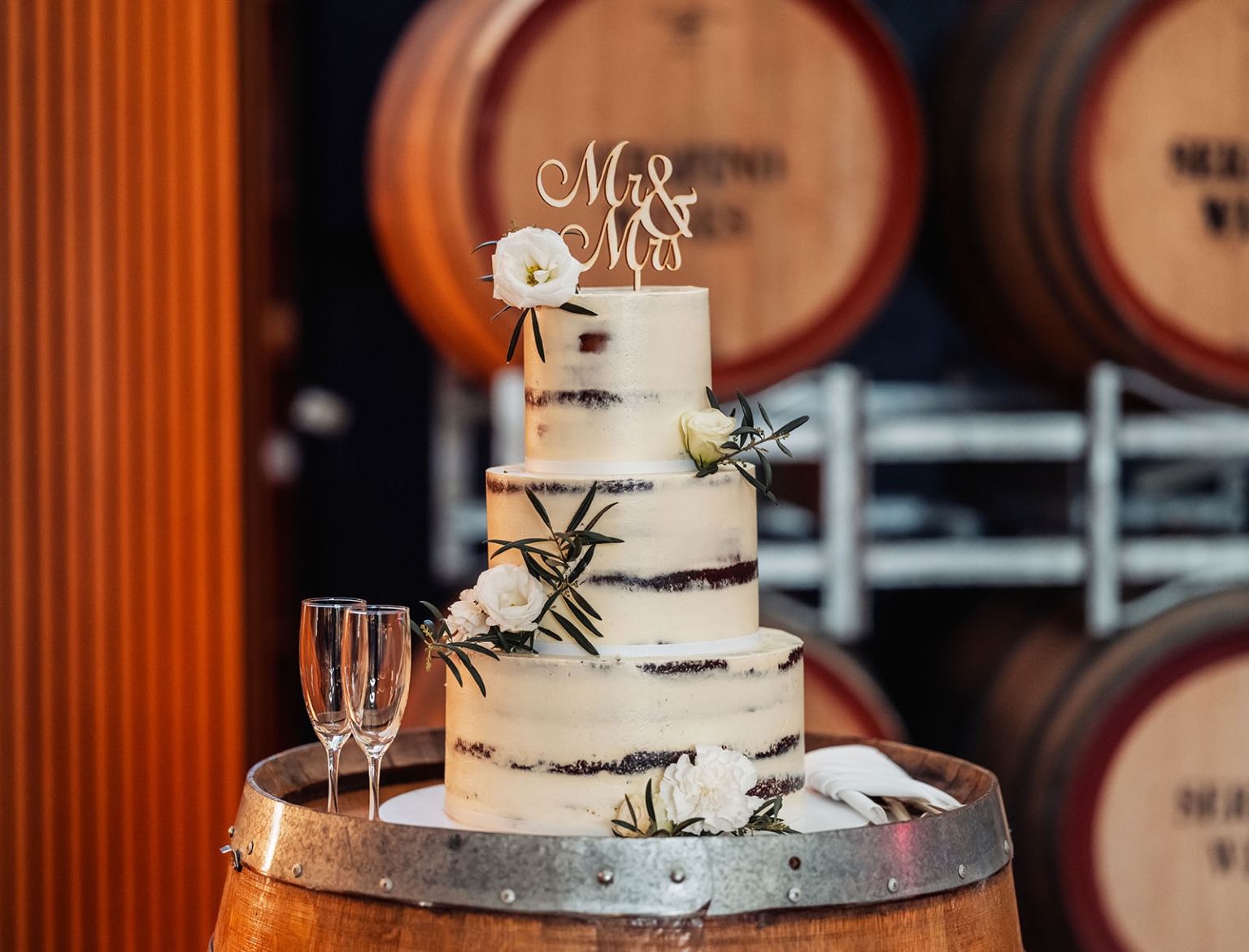 serafino winery wedding – maor & sasha 57 Three-tier semi-naked wedding cake with white flowers and a Mr & Mrs topper, displayed among wine barrels at Serafino Winery.