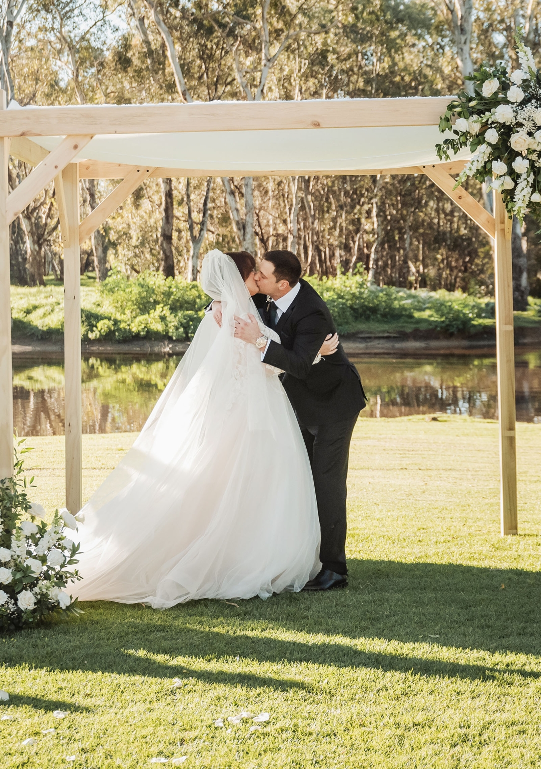 serafino winery wedding – maor & sasha 34 Bride and groom kissing beneath a flower-decorated timber arbour beside a pond at Serafino Winery