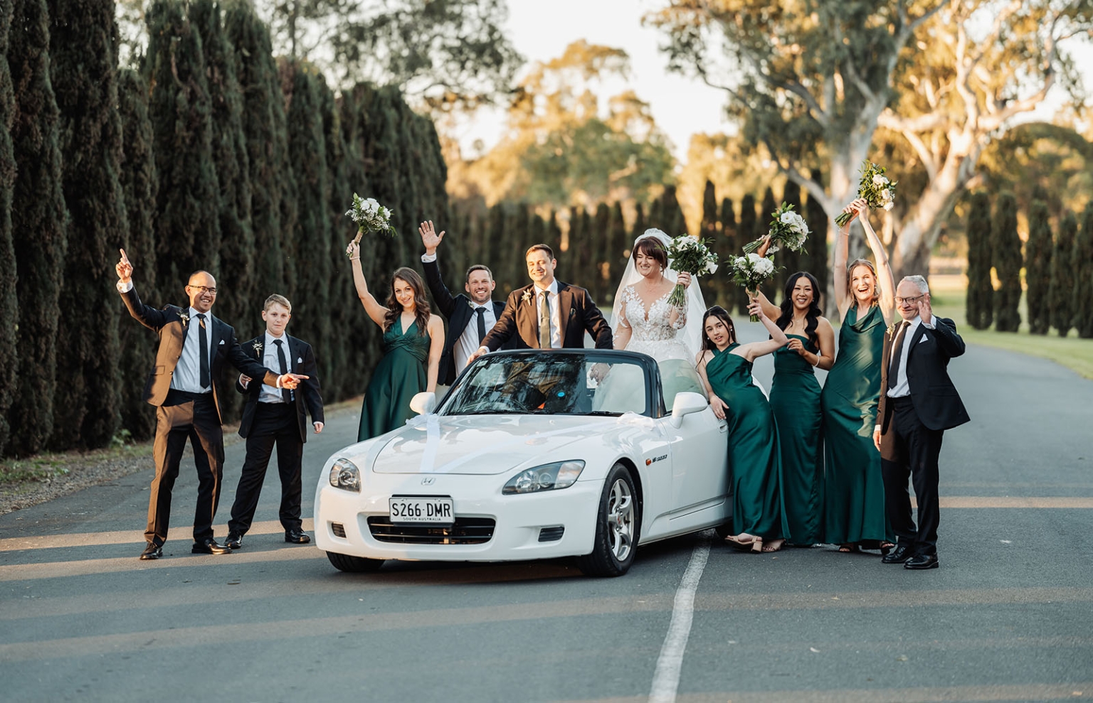 serafino winery wedding – maor & sasha 48 Maor and Sasha with their wedding party beside a white convertible, with bridesmaids raising bouquets on a tree-lined driveway at Serafino Winery