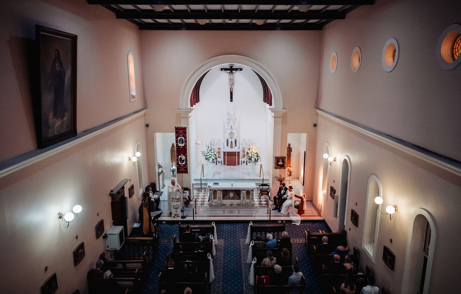 st joseph's chapel wedding – diana & michael 13 Wide elevated view of a wedding ceremony in St Joseph's Chapel, with the couple seated near the altar and guests in pews