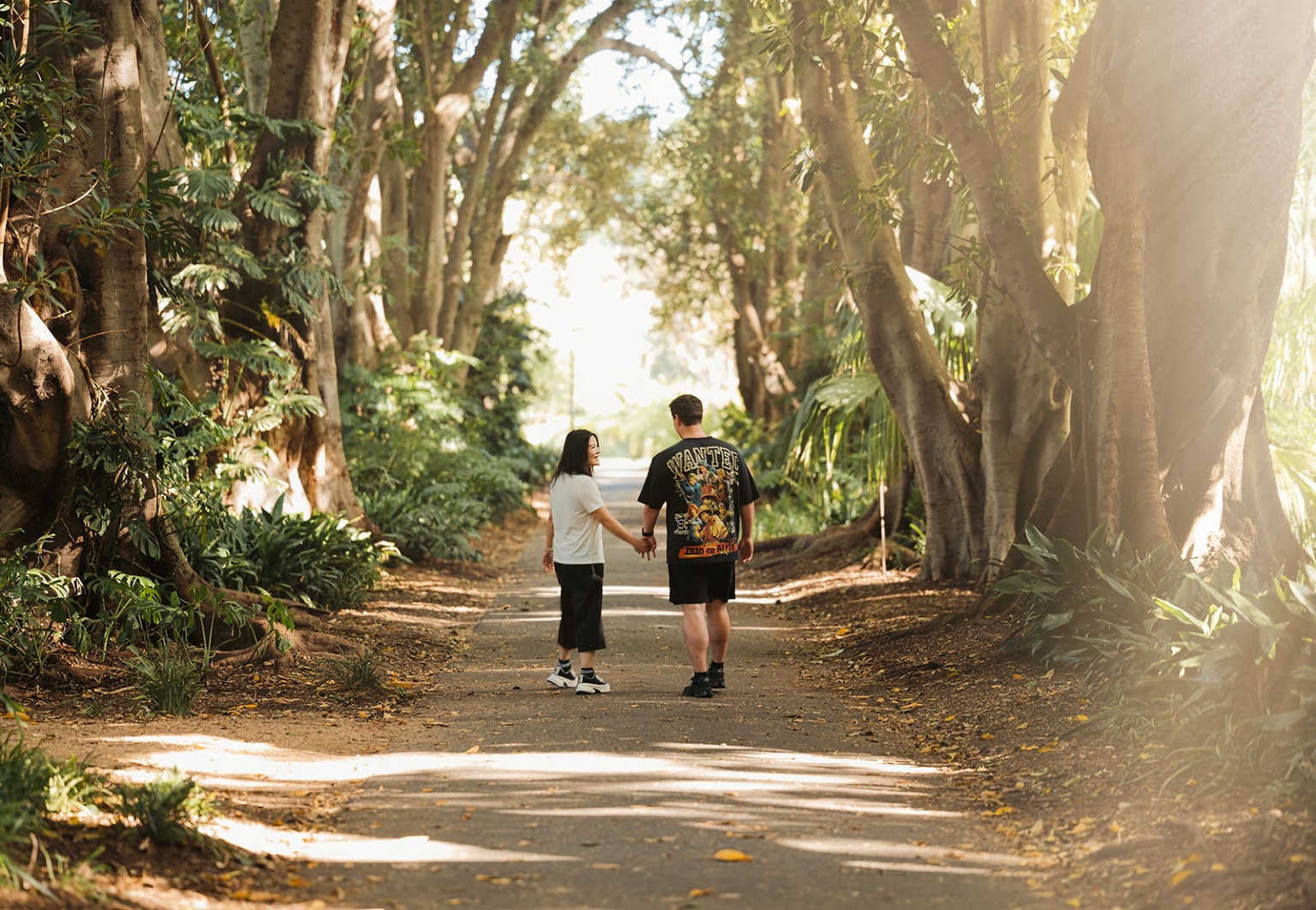 adelaide botanic gardens proposal – stewart & levi 16 Stewart and Levi holding hands on a sunlit, tree-lined path at Adelaide Botanic Gardens