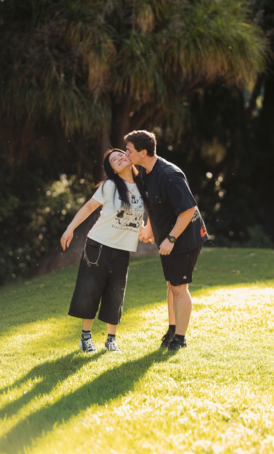 adelaide botanic gardens proposal – stewart & levi 21 Stewart kisses Levi on the cheek as they hold hands on a sunlit lawn at Adelaide Botanic Gardens.