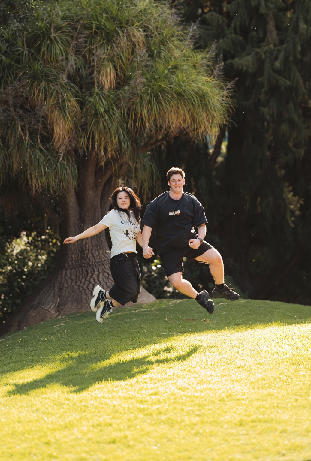 adelaide botanic gardens proposal – stewart & levi 20 Stewart and Levi holding hands and jumping on a sunny lawn at Adelaide Botanic Gardens