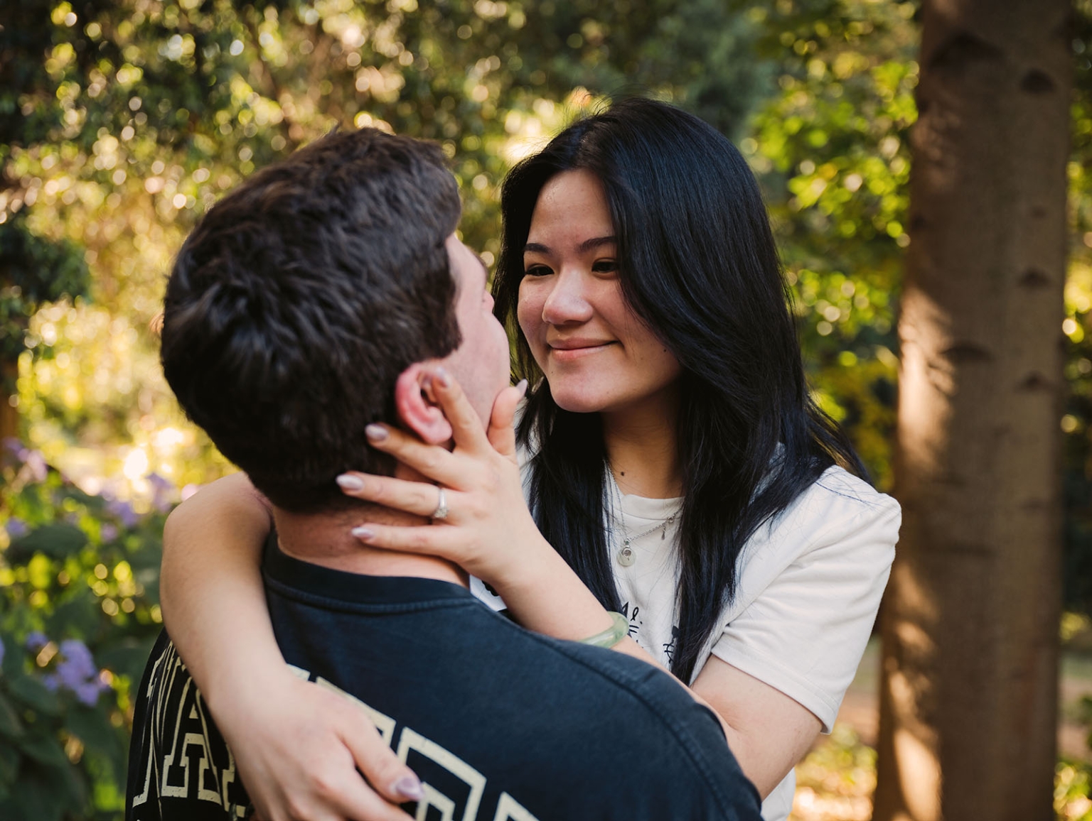 adelaide botanic gardens proposal – stewart & levi 15 Levi embraces Stewart, showing her engagement ring, among sunlit trees at Adelaide Botanic Gardens.