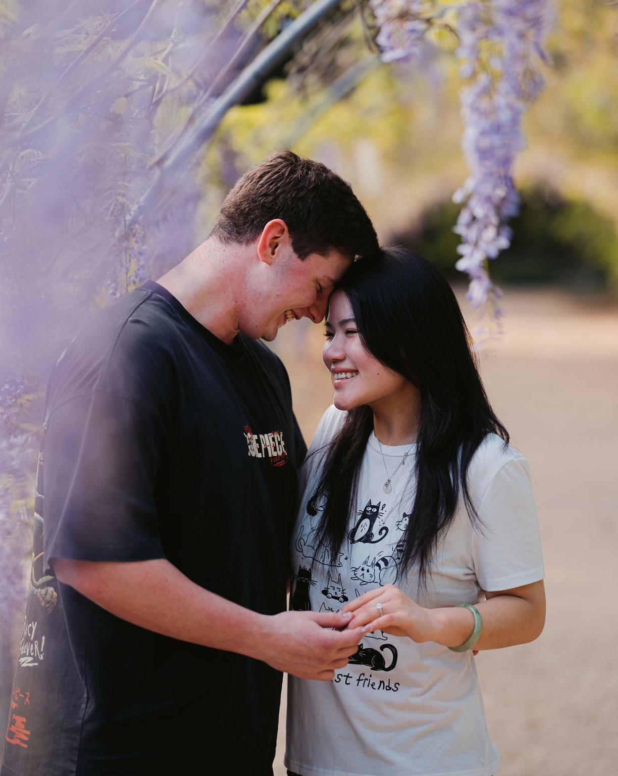 adelaide botanic gardens proposal – stewart & levi 13 Stewart and Levi smiling together as he holds her hand to show her engagement ring beneath purple flowers at Adelaide Botanic Gardens