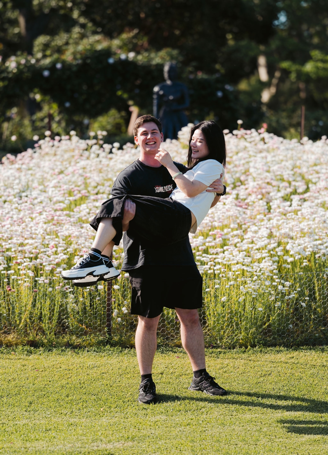 adelaide botanic gardens proposal – stewart & levi 5 Stewart carries Levi as they smile beside a bed of pale roses at Adelaide Botanic Gardens.