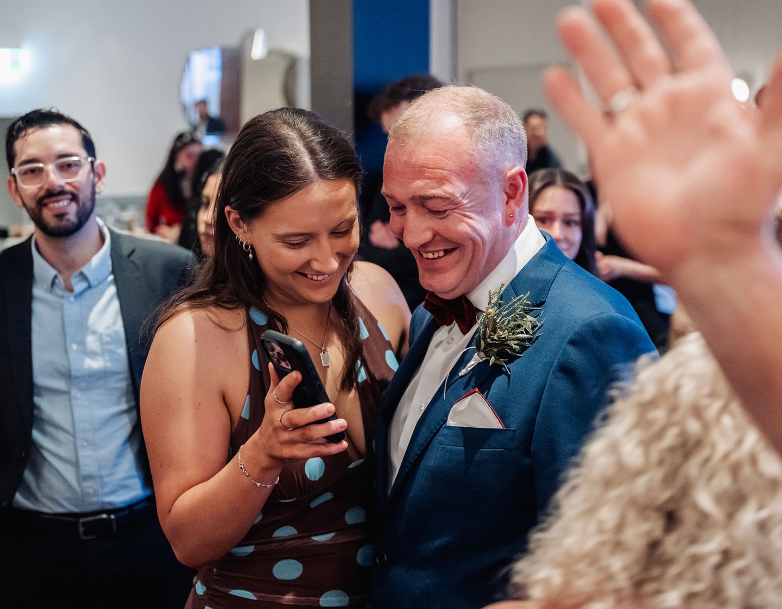hotel royal wedding - brad & manuel 31 Wedding guest holding a phone beside a smiling groom in a blue suit, surrounded by guests indoors