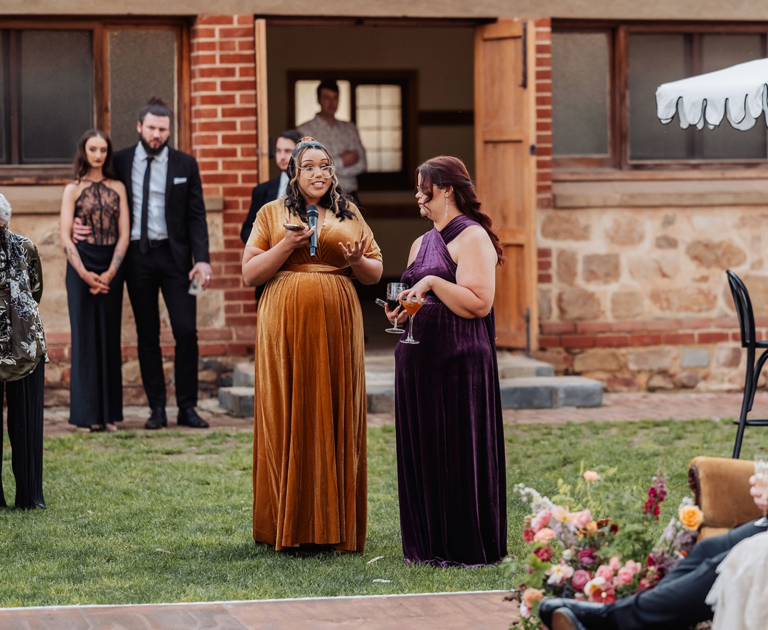 burbrook wedding – amber & angus | kuitpo forest wedding 54 Wedding guest in a gold velvet dress giving a speech with a microphone beside a woman holding a drink on a lawn
