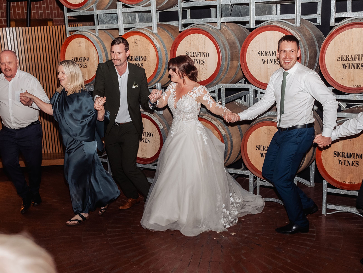 serafino winery wedding – maor & sasha 64 Bride and wedding guests dance hand in hand in front of Serafino Wines barrels.
