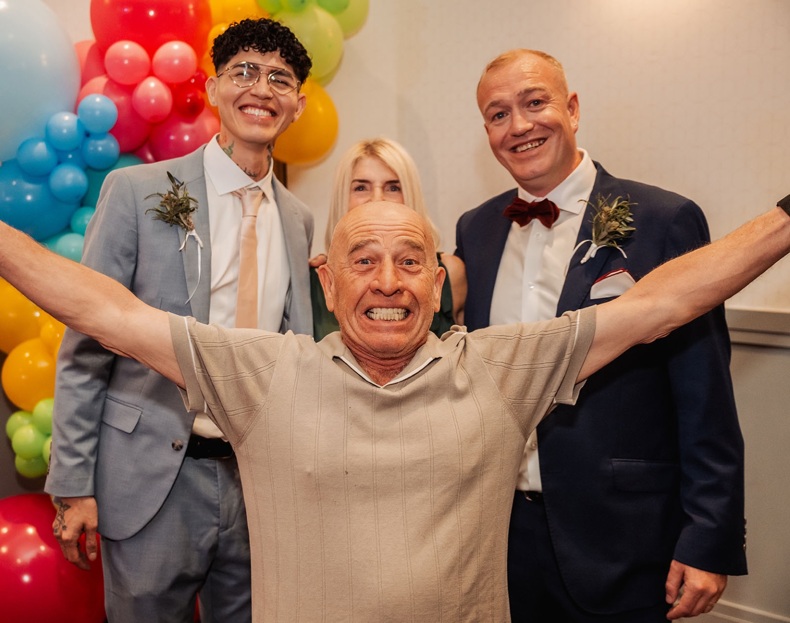 hotel royal wedding - brad & manuel 33 Smiling wedding guests pose in front of a colourful rainbow balloon display, with one man raising his arms in the foreground.