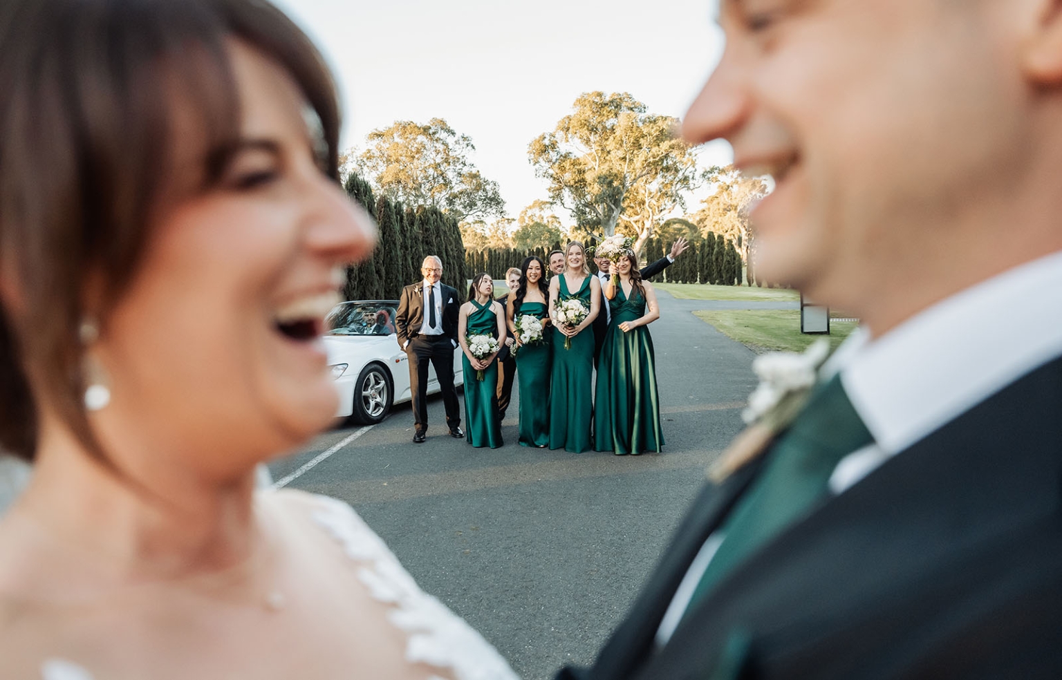 serafino winery wedding – maor & sasha 47 Laughing bride and groom in the foreground with their wedding party in green dresses posing behind them on a tree-lined driveway.