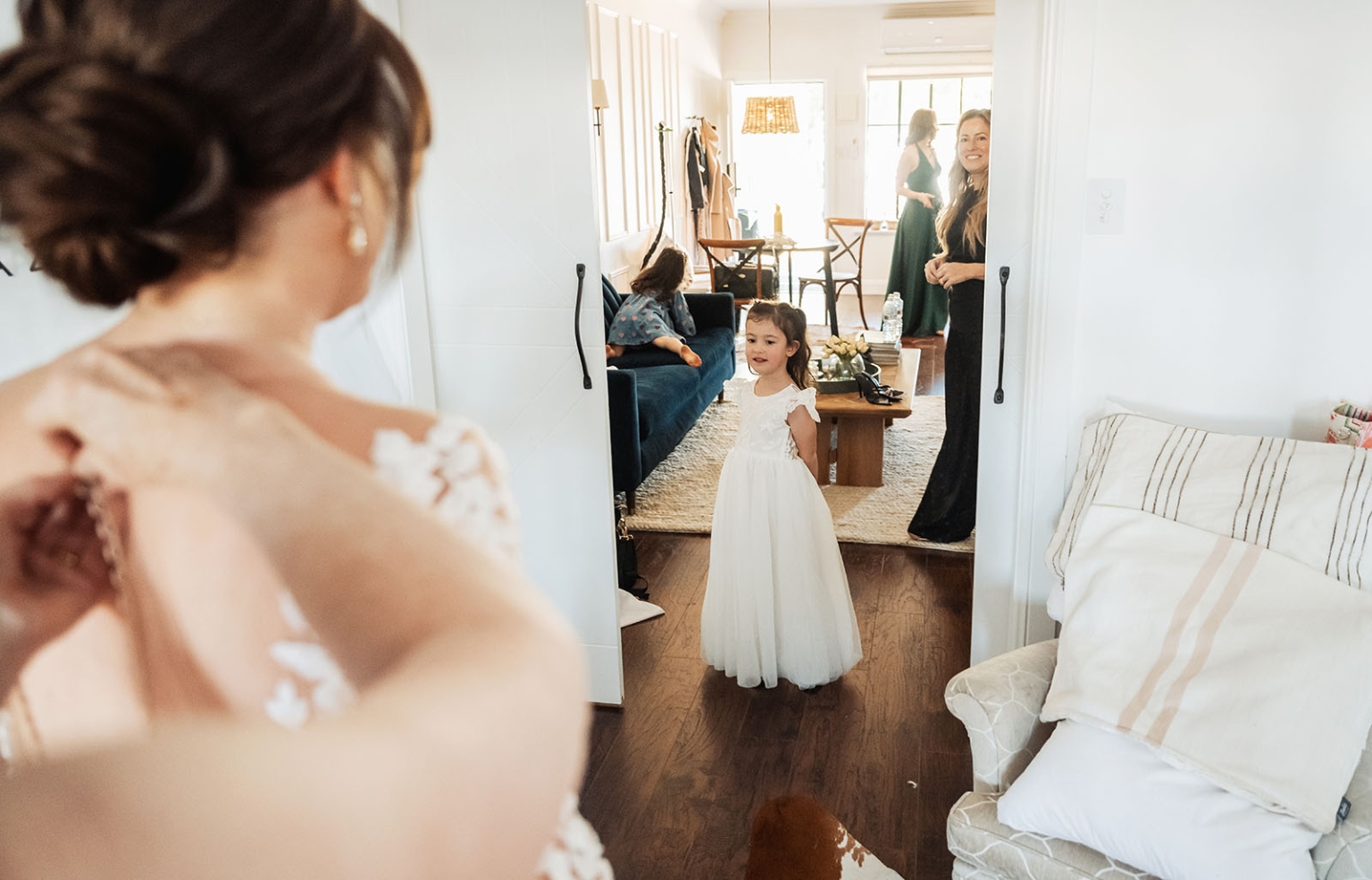 serafino winery wedding – maor & sasha 19 Flower girl in a white dress watches as the bride prepares in a bright apartment, with wedding guests in the adjoining room.