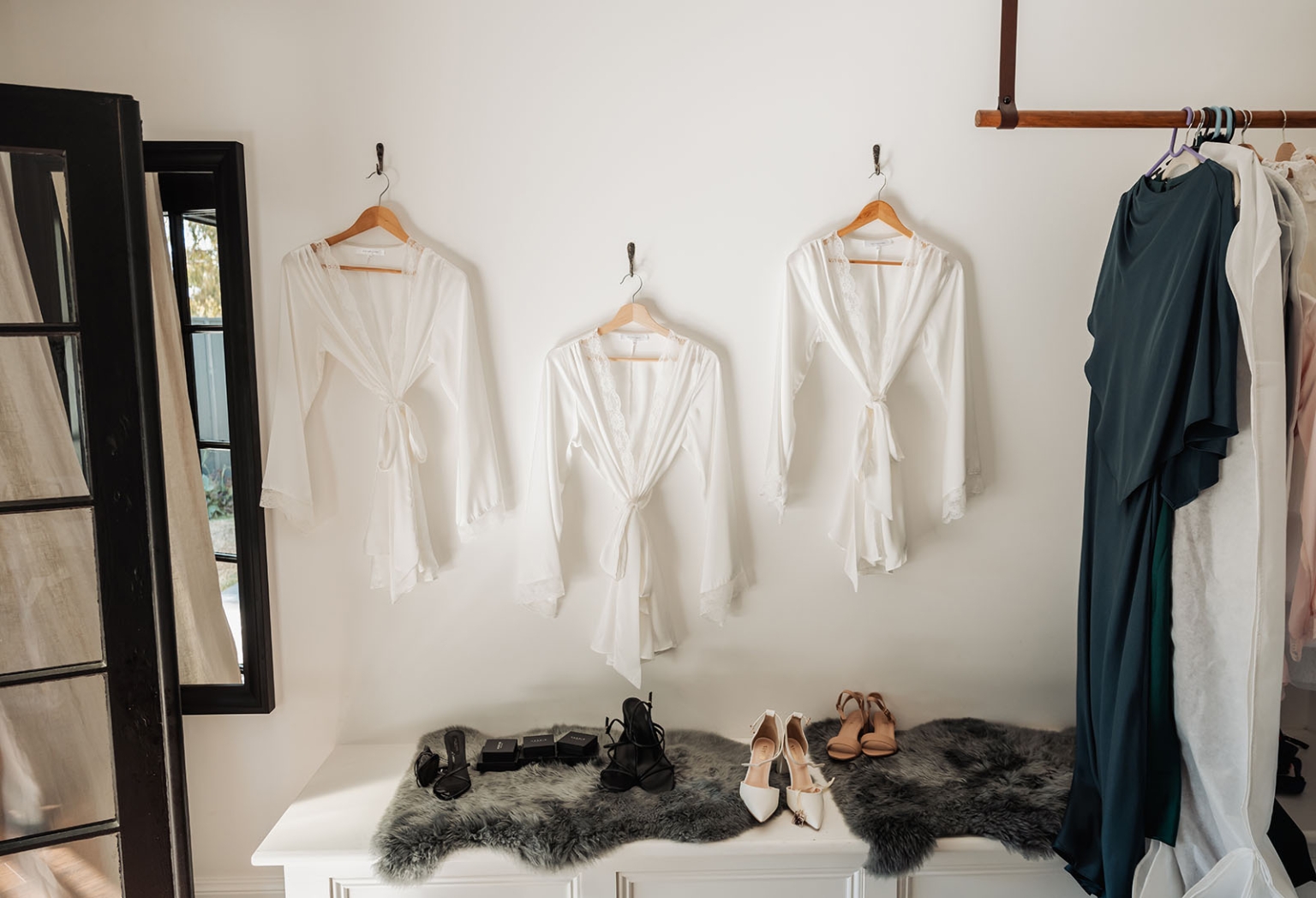 serafino winery wedding – maor & sasha 15 Three white satin robes hanging above a bench with wedding shoes, alongside formal dresses on a clothes rack