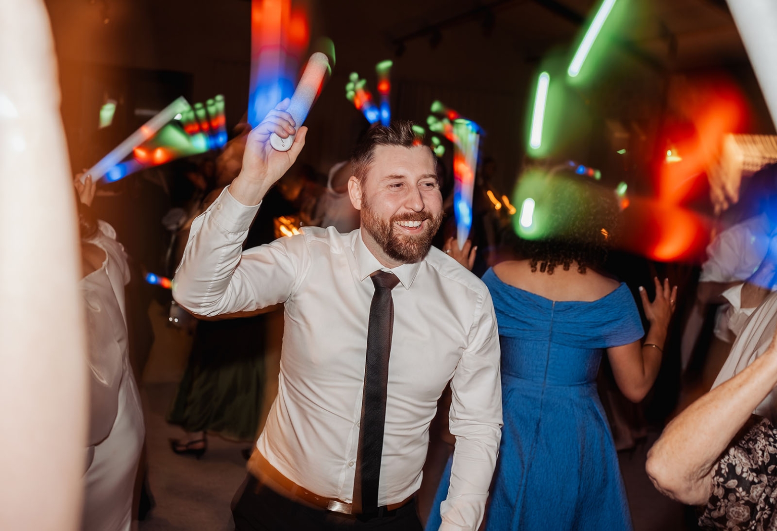 st joseph's chapel wedding – diana & michael 57 Man in a white shirt and tie dancing with a glowing foam stick among guests under colourful party lights