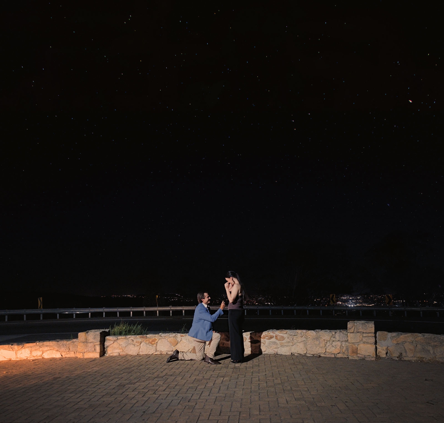 Josh kneels to propose to Kelly beneath a star-filled sky overlooking Adelaide city lights at Windy Point Lookout.