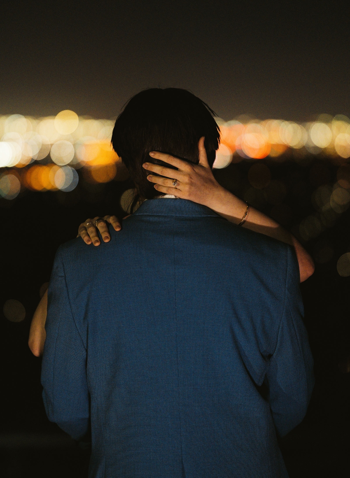 Kelly embraces Josh, her engagement ring visible against his shoulder, with Adelaide city lights glowing behind them at night.