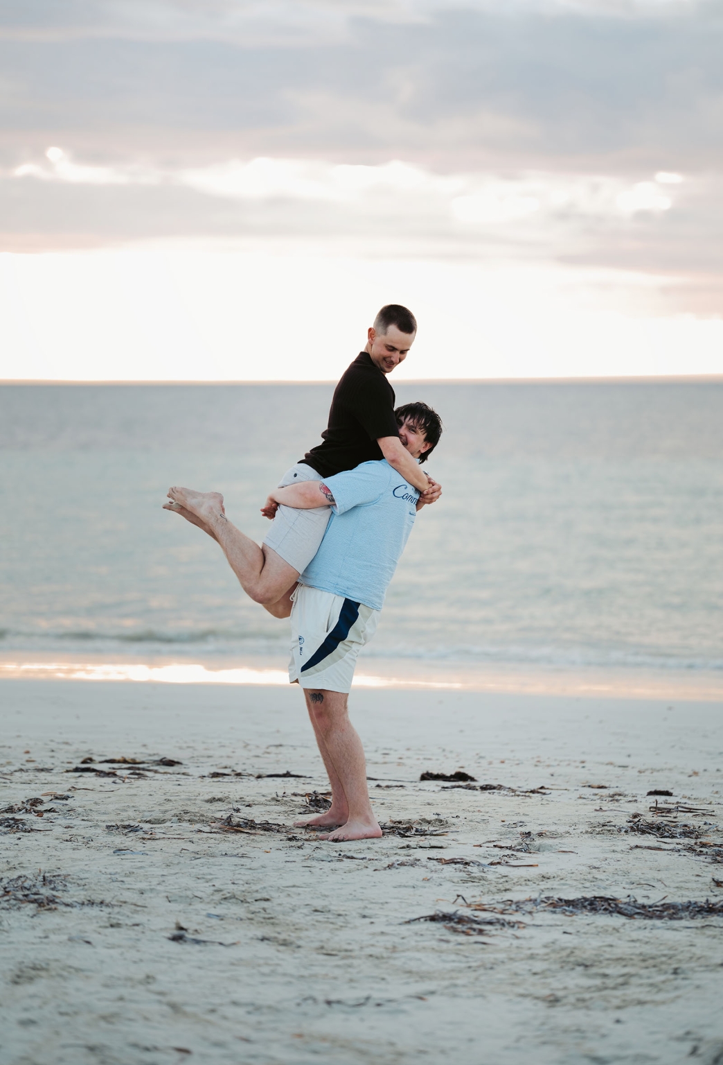 moonta's simms cove proposal – zoe & nathan 24 Nathan lifts Zoe in an embrace on the beach at Simms Cove, with calm water and a cloudy sunset behind them.