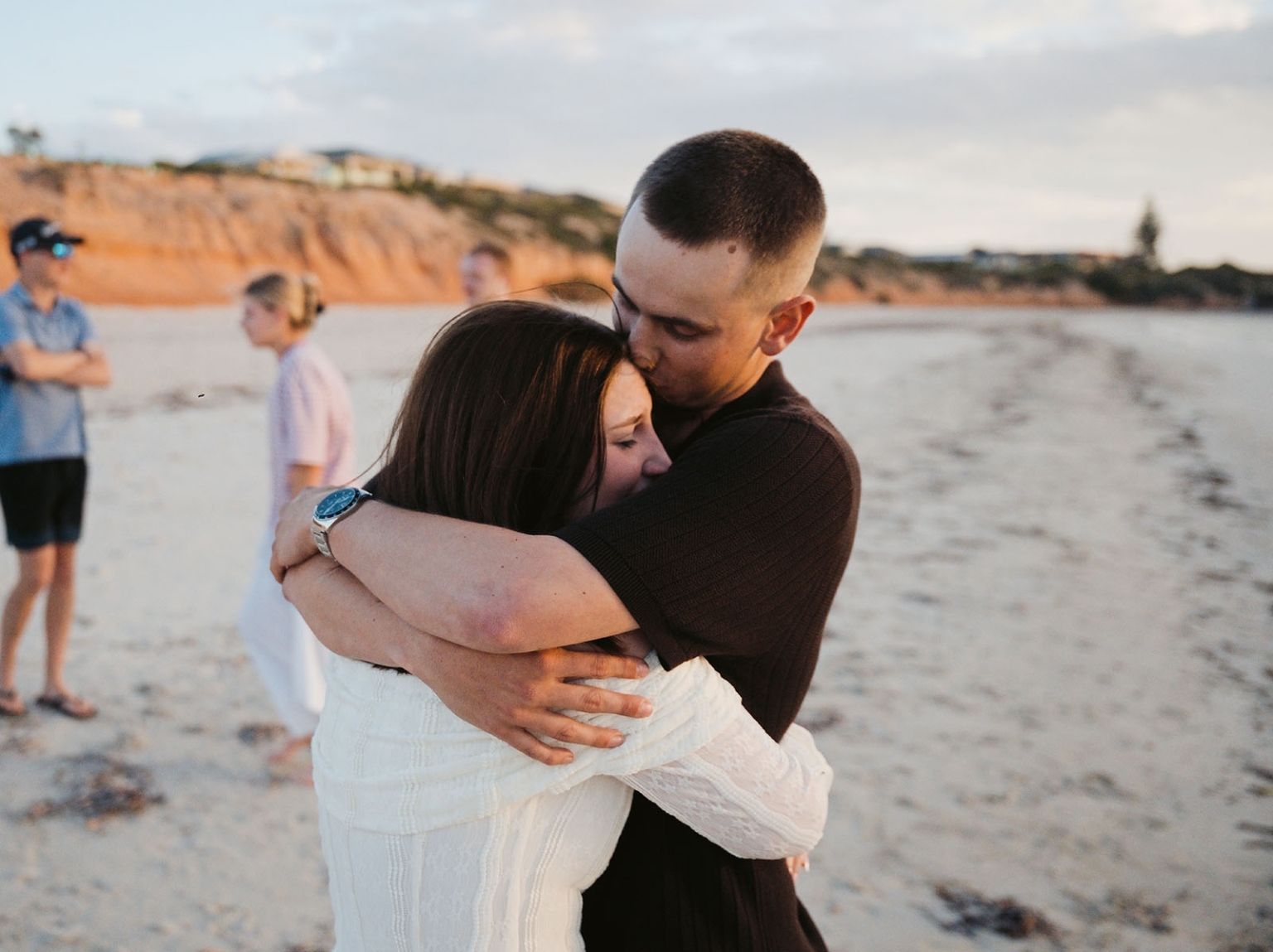 moonta's simms cove proposal – zoe & nathan 7 Zoe and Nathan embracing on the sand at Simms Cove, with friends standing nearby