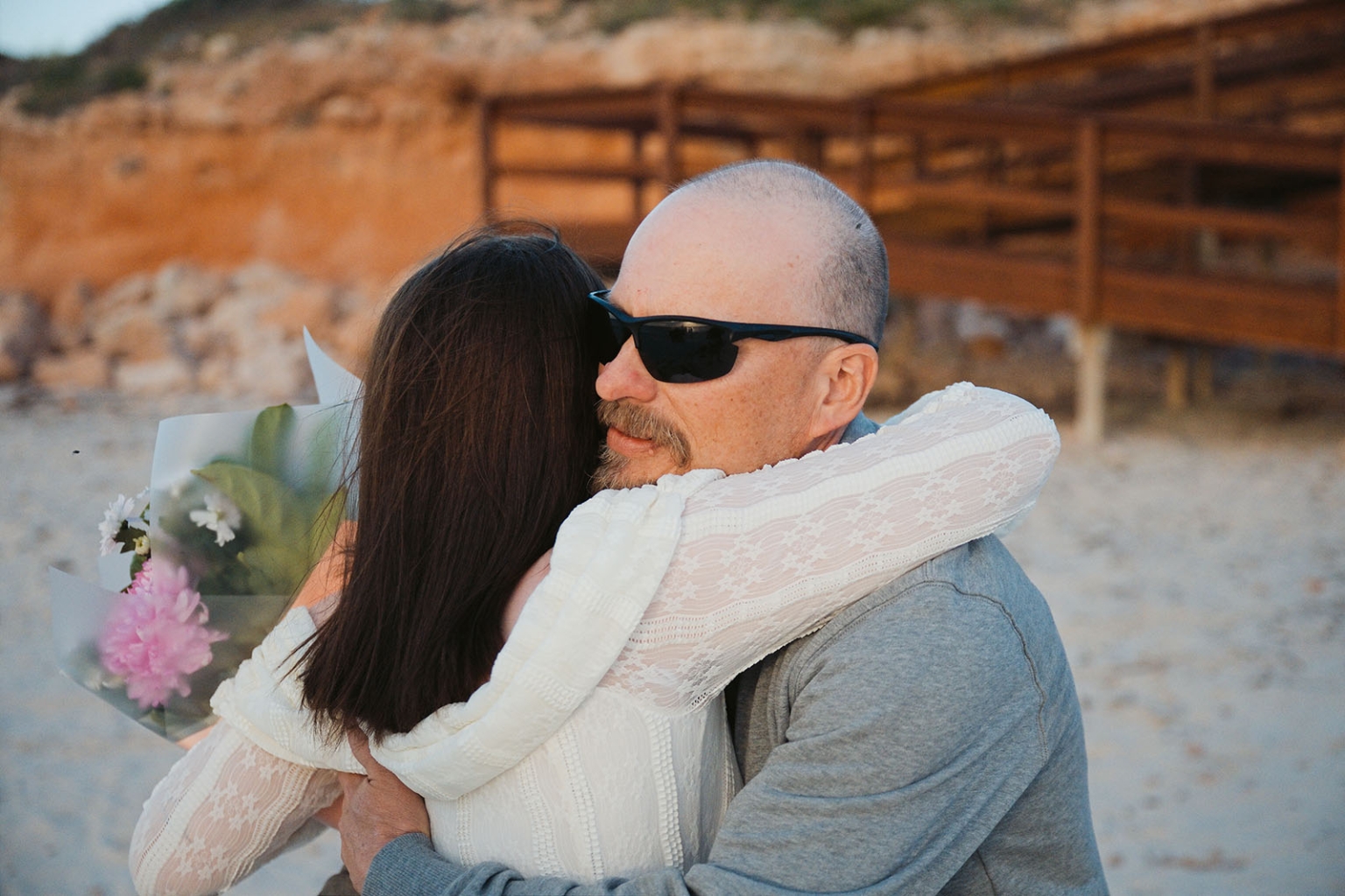 moonta's simms cove proposal – zoe & nathan 11 Nathan and Zoe embracing on Simms Cove beach, with Zoe holding a bouquet of flowers.