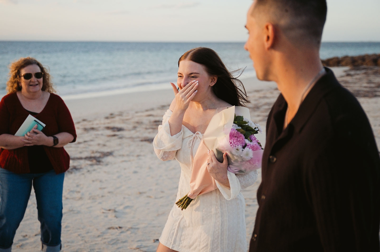 moonta's simms cove proposal – zoe & nathan 12 Zoe covering her mouth in surprise while holding a bouquet beside Nathan on Simms Cove beach, with a smiling guest nearby